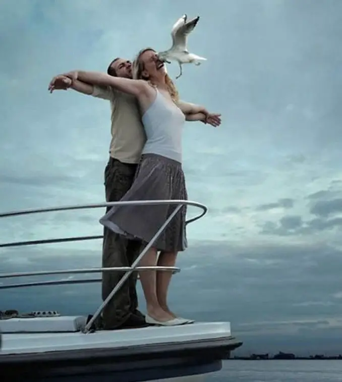 A couple standing at the front of a boat and a seagull has flown into the woman's face.