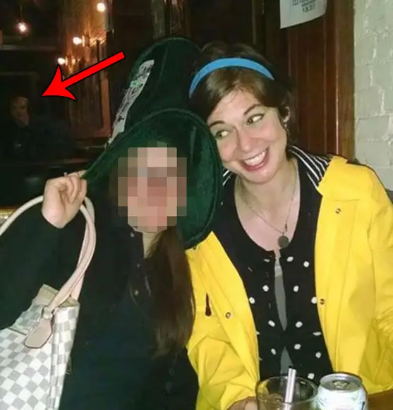 Two friends pose for a photo in a pub. A scary face can be seen in the background. Is it a ghost?